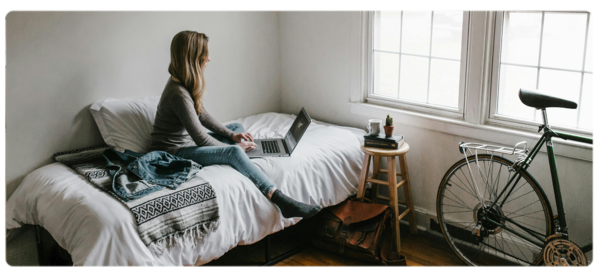 A student in a foreign country sits on a dorm bed and does homeowork on a laptop. In the corner, a bicycle is stored.