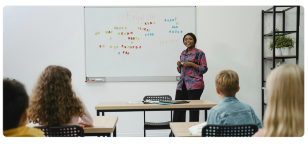 A young teacher stands at a marker board at the front of a class of students.