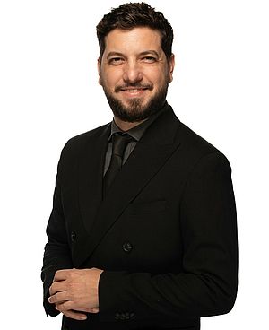 Tiago Arrais stands in front of a white background and smiles for a photo.