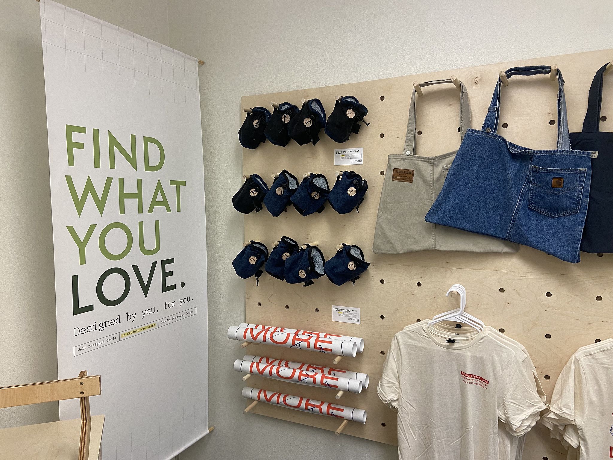 Tshirts, tote bags, posters, and chalk bags hang on a wooden display next to a Well-Designed Goods sign.