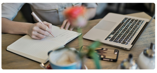 A student sits in front of a laptop next to a cup of coffee and writes in a journal.