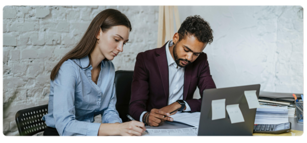 Two people sit at a desk covered in papers engaged in an editing process.