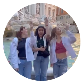 Three students throw coins over their shoulder into a fountain