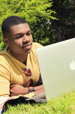 Male student works on his laptop while laying on the grass.