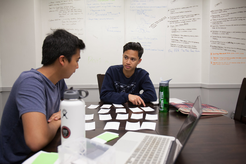two students studying at a table with a whiteboard behind