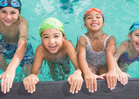 Smiling kids in swim caps and goggles lined up in the water along the edge of a pool.